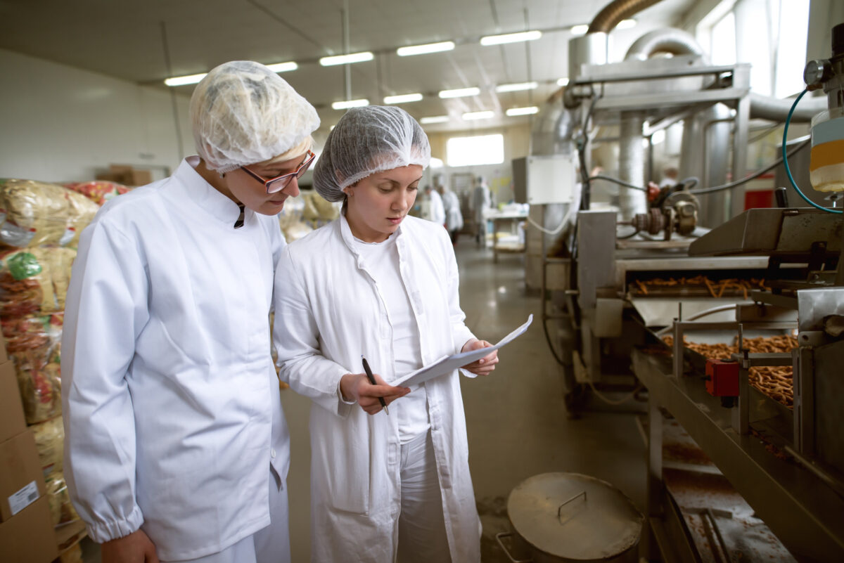 Deux ouvrières contrôlent la qualité des aliments dans une usine alimentaire. L'une d'elles tient des documents et un stylo, l'autre regarde des documents.