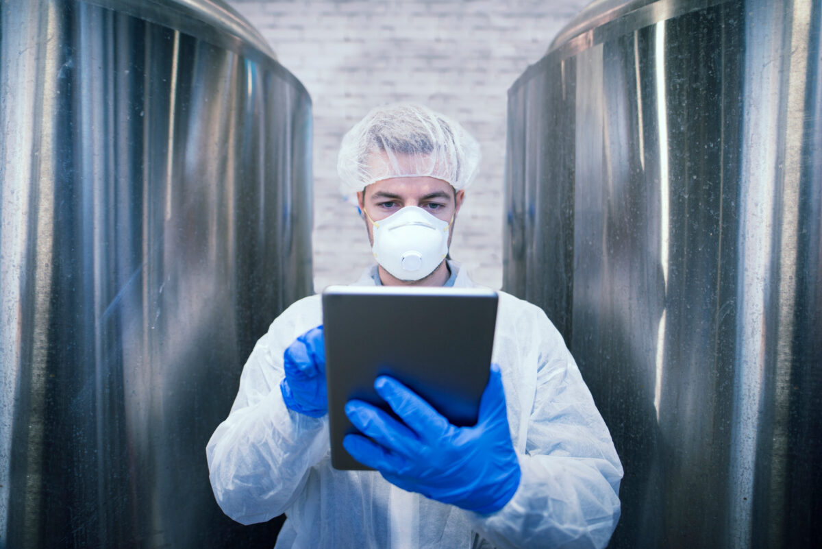 Portrait d'un technologue en uniforme de protection blanc tenant une tablette dans une usine de production alimentaire. Travailleur industriel avec filet à cheveux et masque contrôlant la production dans l'industrie chimique ou pharmaceutique.