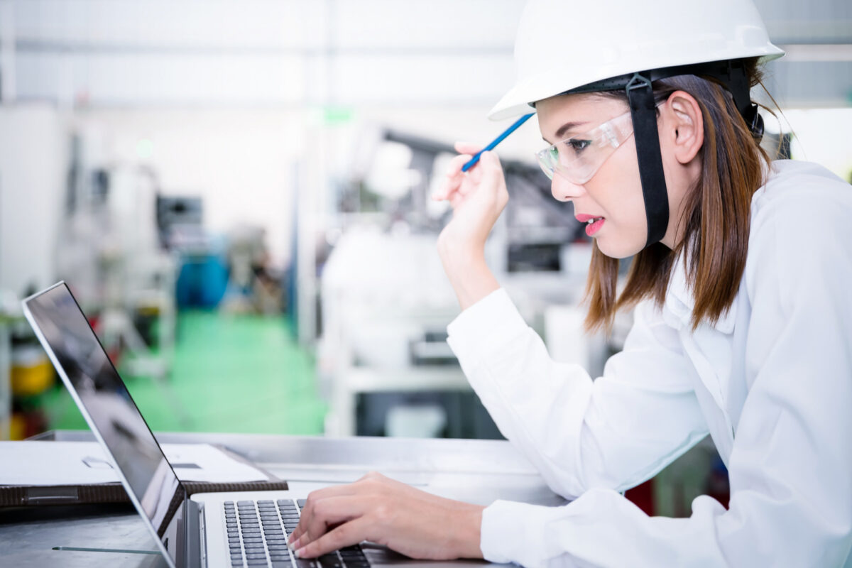 Une ingénieure industrielle coiffée d'un casque utilise un ordinateur portable dans une usine.